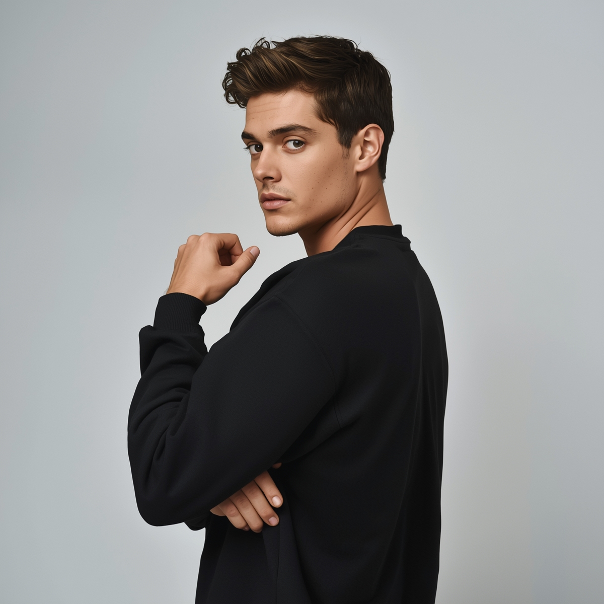 Male model standing in a relaxed pose, body turned slightly to the side with head turned toward the camera. One arm bent, hand resting near the chin or jawline, and the other arm loosely folded across the body. Expression is calm and confident. Simple studio background with soft lighting